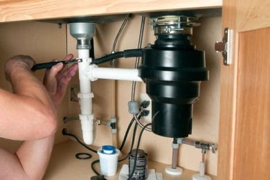 Plumber repairing a garbage disposal beneath a kitchen sink in Greenfield